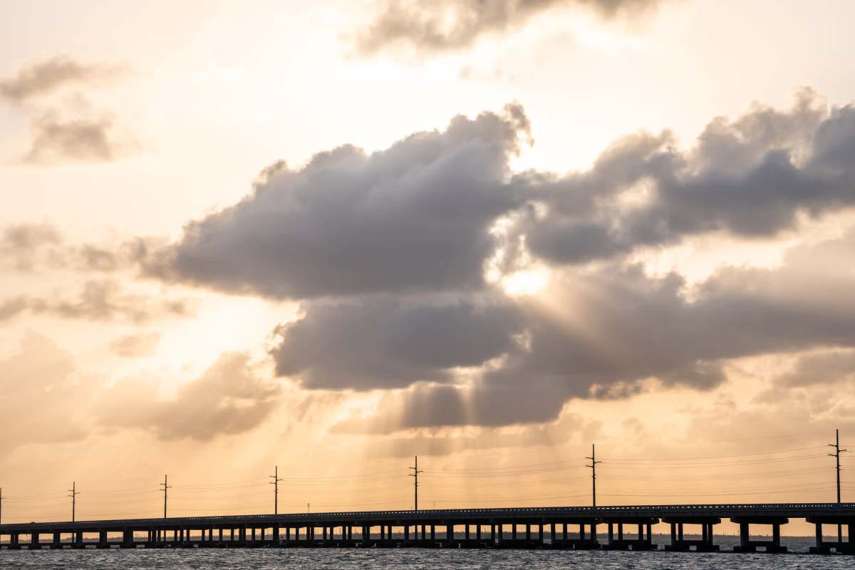 A long bridge stretches over water with dramatic clouds and rays of sunlight breaking through the sky.
