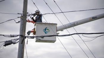 A worker in a lift repairs power lines on a utility pole under a cloudy sky.