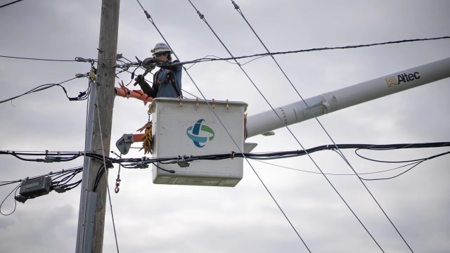 A worker in a lift repairs power lines on a utility pole under a cloudy sky.