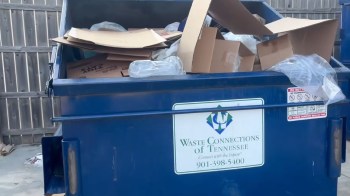 A blue dumpster filled with cardboard boxes and plastic bags, located next to a wooden fence.