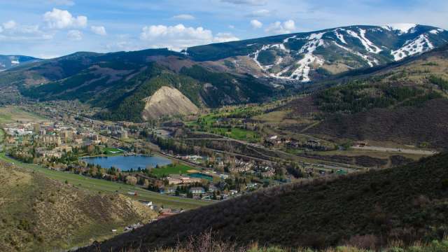 A panoramic view of a mountainous landscape with a valley, a lake, and a small town below under a blue sky.