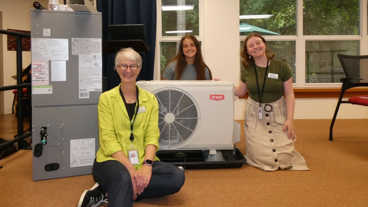 Three people pose next to a heat pump unit.