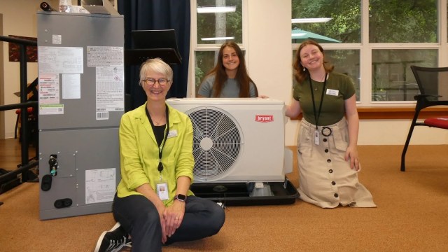 Three people pose next to a heat pump unit.