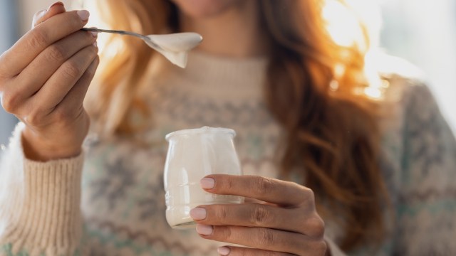 A person holding a small jar of yogurt and a spoon, wearing a cozy knit sweater.