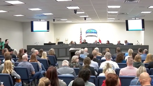 A crowded meeting room with attendees facing a panel of officials and screens displaying information.
