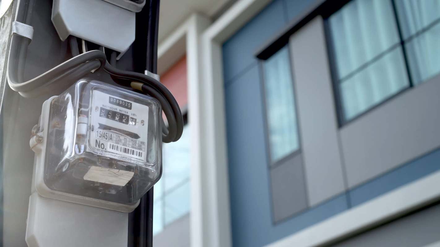 An electric meter attached to a building with modern architecture in the background.
