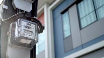 An electric meter attached to a building with modern architecture in the background.