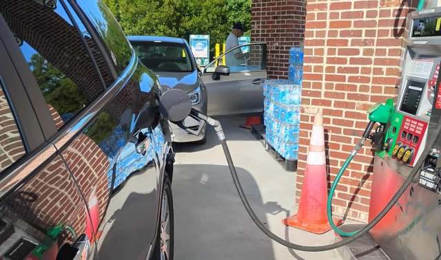 A car at a gas station with a pump nozzle inserted, alongside stacked water bottles and a person at the entrance.