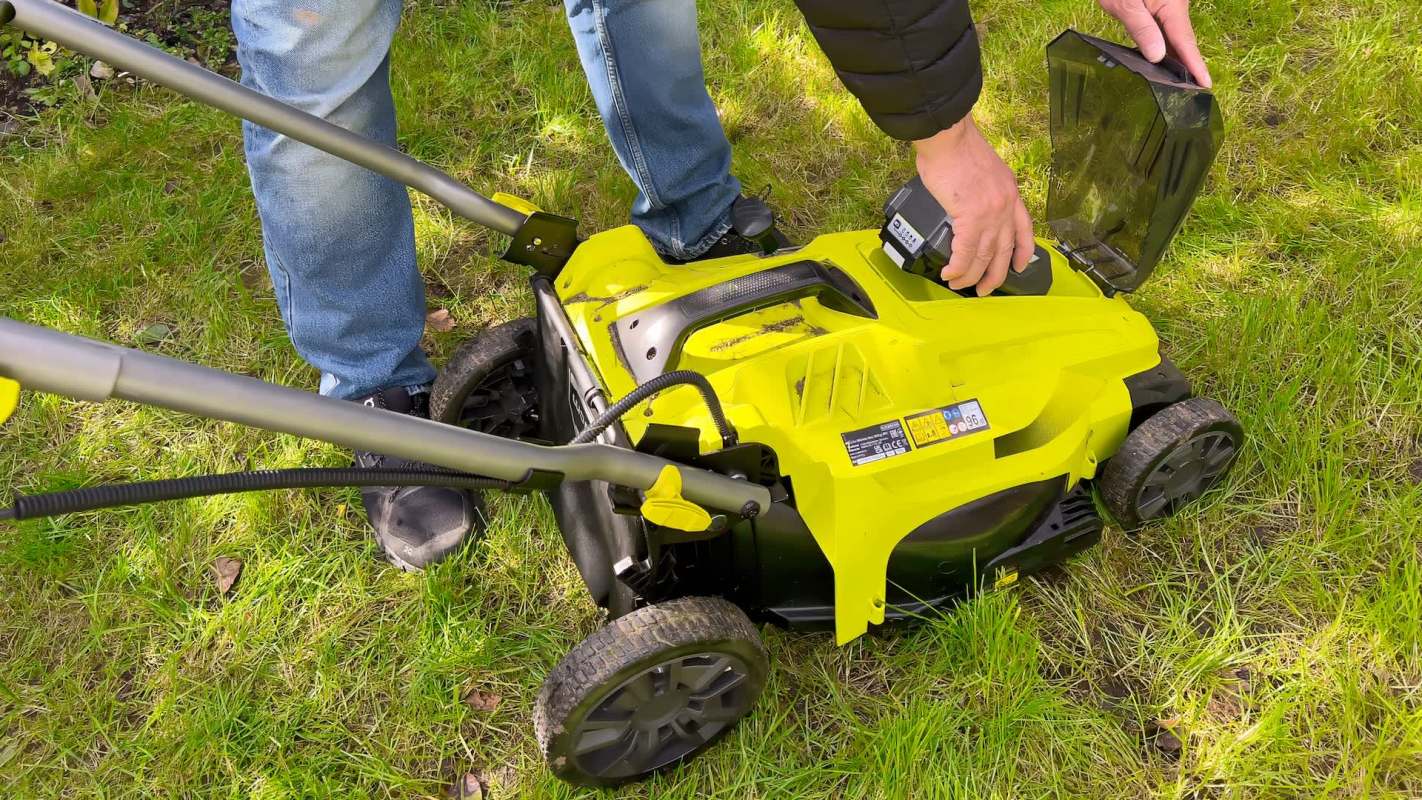 A person is attaching a battery to a green and black electric lawn mower on a grassy area.