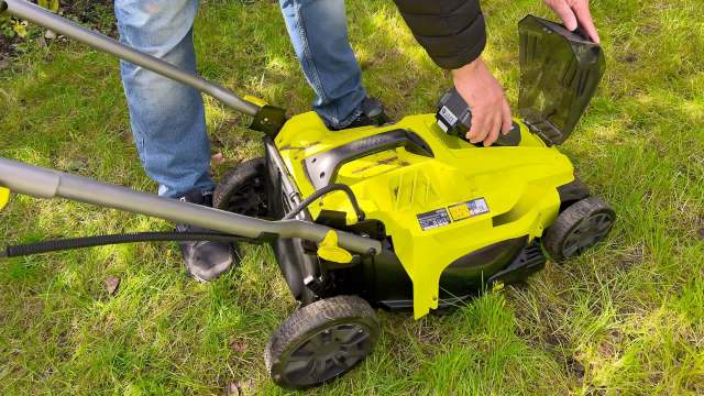 A person is attaching a battery to a green and black electric lawn mower on a grassy area.