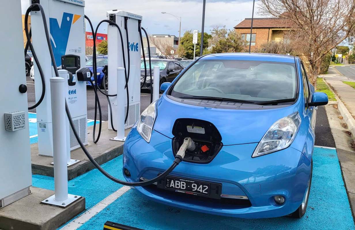 A blue electric car is charging at a public charging station with two chargers visible in the background.
