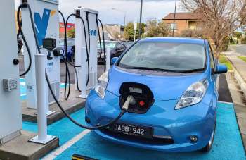 A blue electric car is charging at a public charging station with two chargers visible in the background.