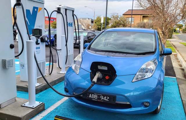 A blue electric car is charging at a public charging station with two chargers visible in the background.