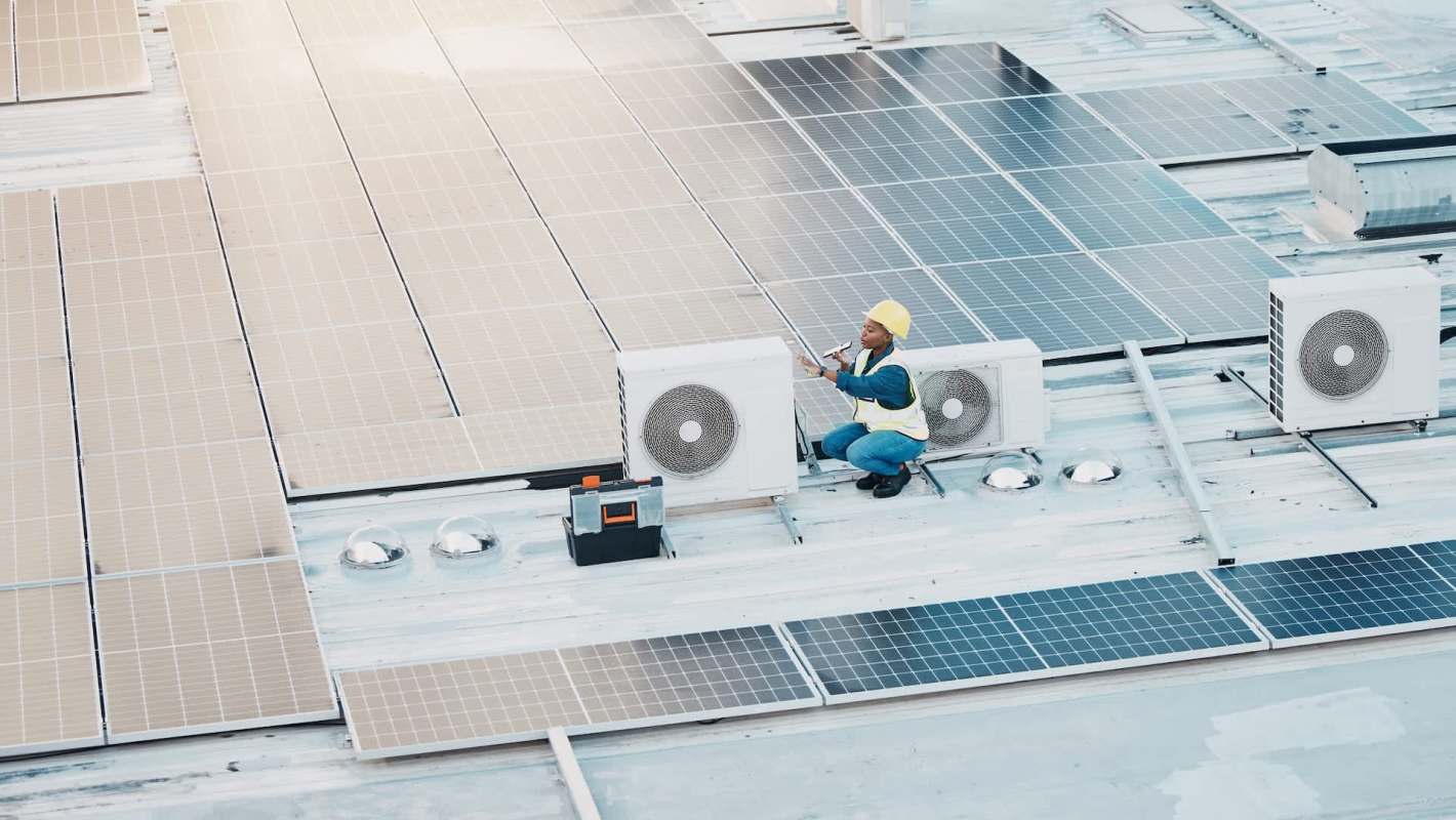 A worker in a hard hat and safety vest inspects air conditioning units on a rooftop covered with solar panels.