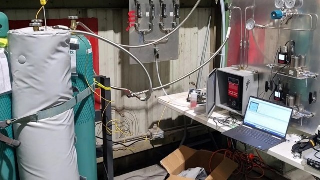 A laboratory setup featuring gas cylinders, tubing, gauges, and a computer on a workbench.