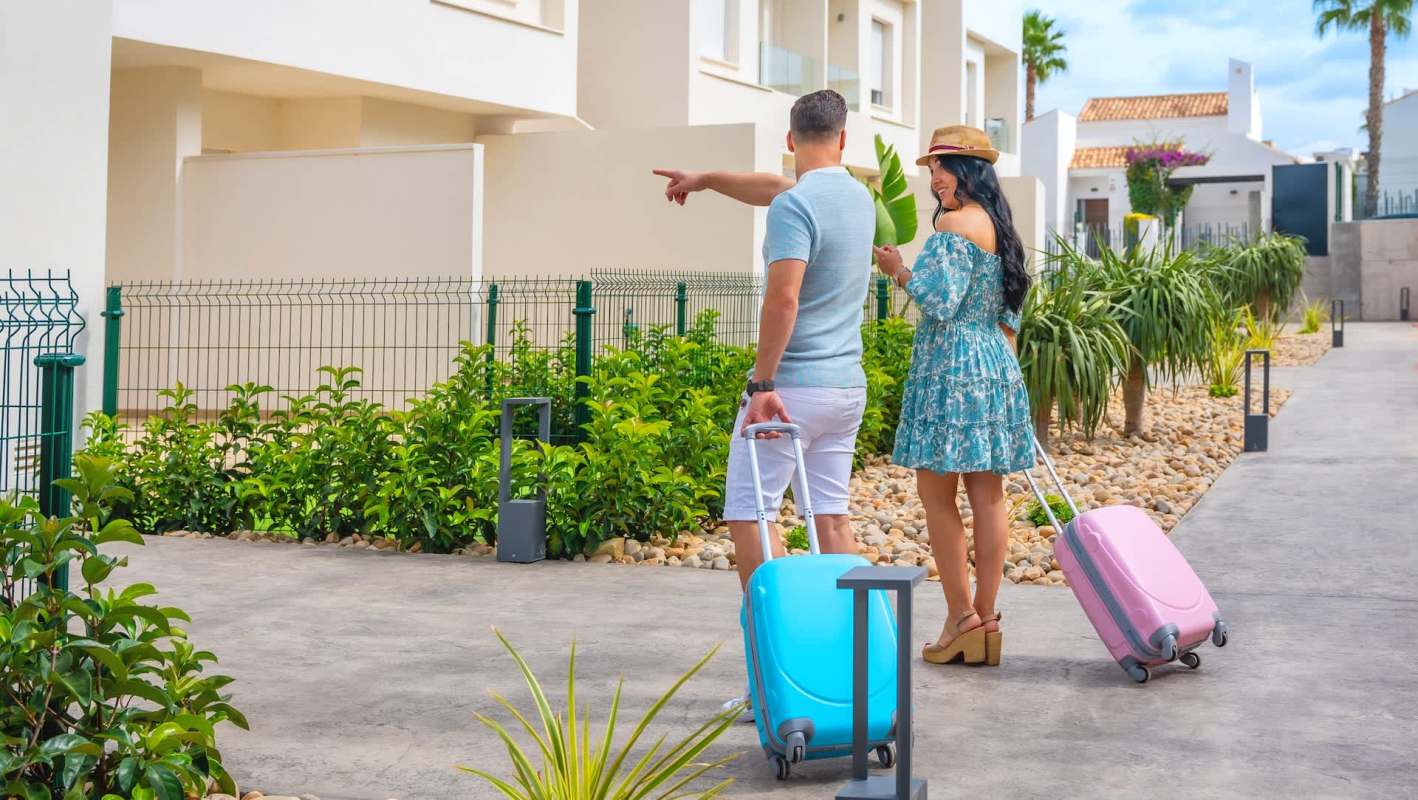 A couple with suitcases enjoys a sunny day in a garden, pointing at a nearby building.