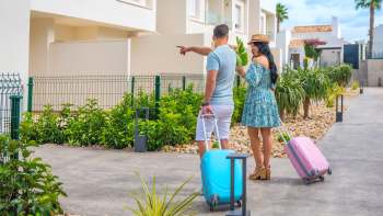 A couple with suitcases enjoys a sunny day in a garden, pointing at a nearby building.