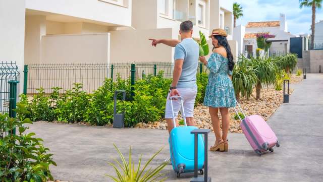 A couple with suitcases enjoys a sunny day in a garden, pointing at a nearby building.