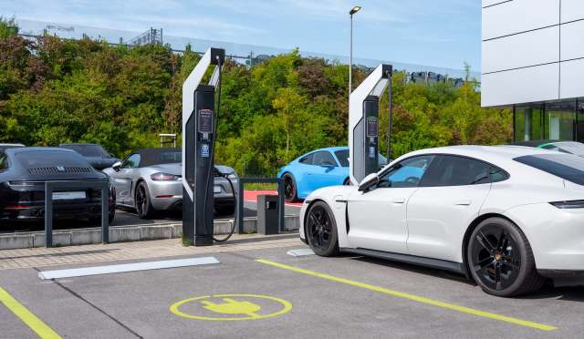 A white electric car is charging at a station in a parking lot with several other cars nearby.