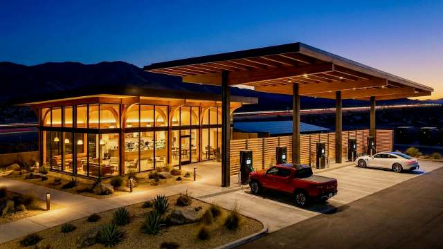 A modern electric vehicle charging station with a sleek wooden structure and illuminated lounge area at dusk.