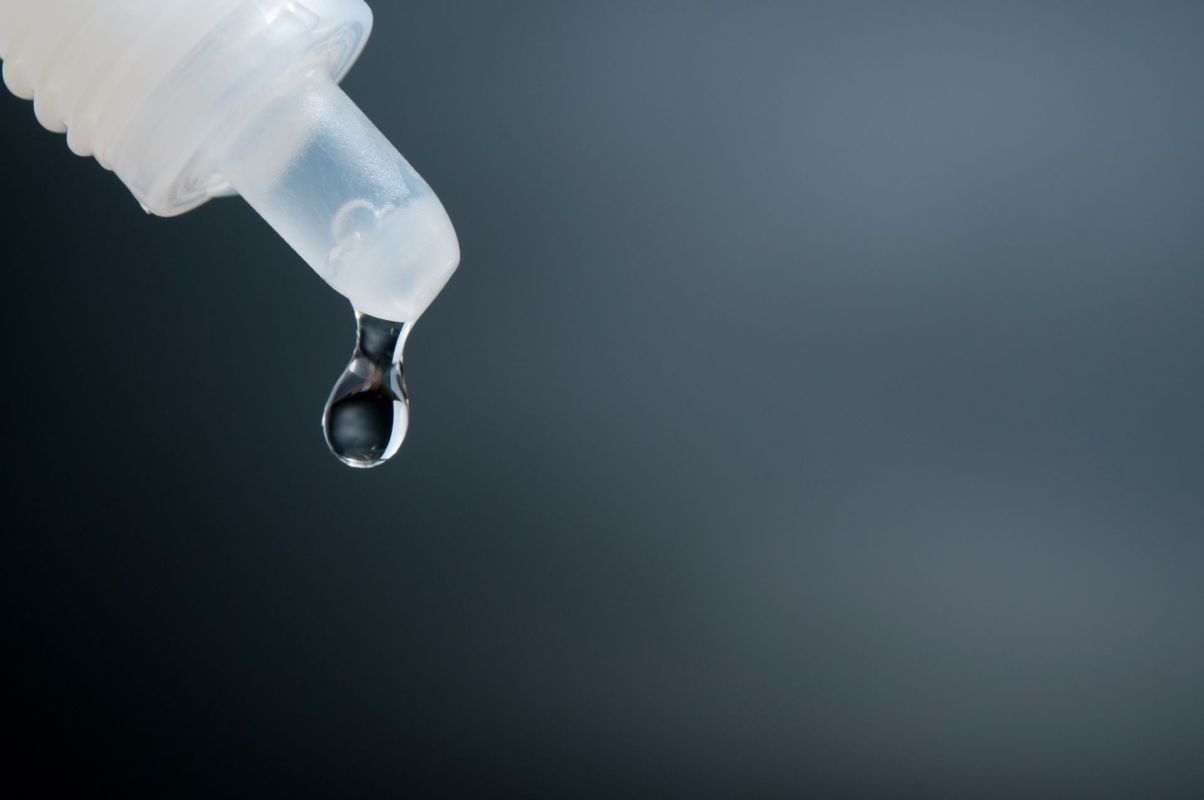A clear drop of liquid is suspended from the tip of a eye dropper against a dark background.