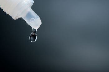 A clear drop of liquid is suspended from the tip of a eye dropper against a dark background.