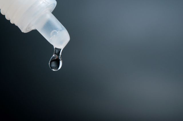 A clear drop of liquid is suspended from the tip of a eye dropper against a dark background.