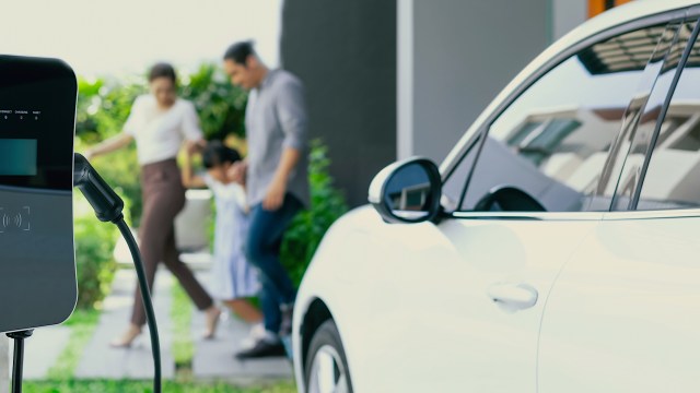 A family walks together near a white electric vehicle charging station in a landscaped outdoor setting.