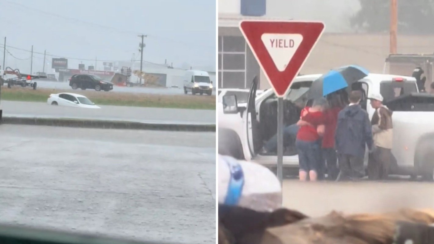 A flooded road with a partially submerged car and a group of people during a rescue.