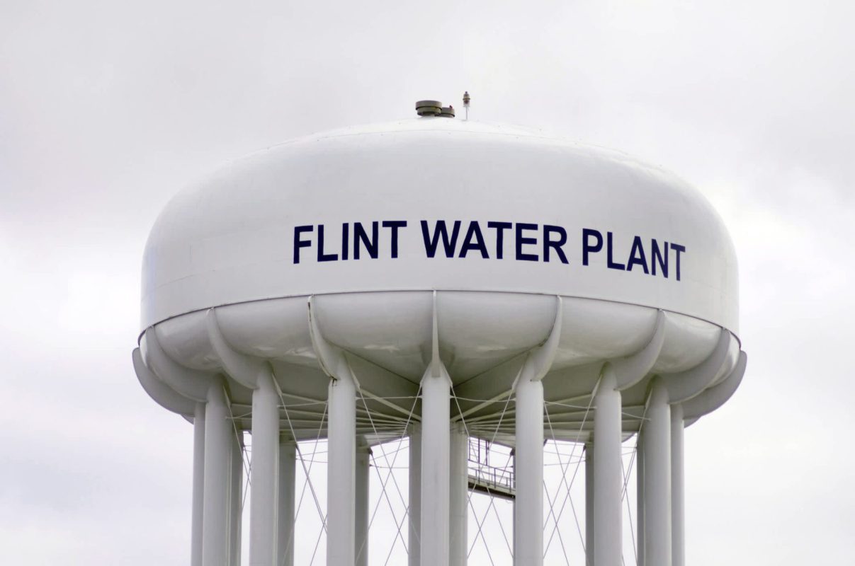 A close-up of a large white water tower labeled "FLINT WATER PLANT" against a cloudy sky.
