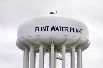 A close-up of a large white water tower labeled "FLINT WATER PLANT" against a cloudy sky.