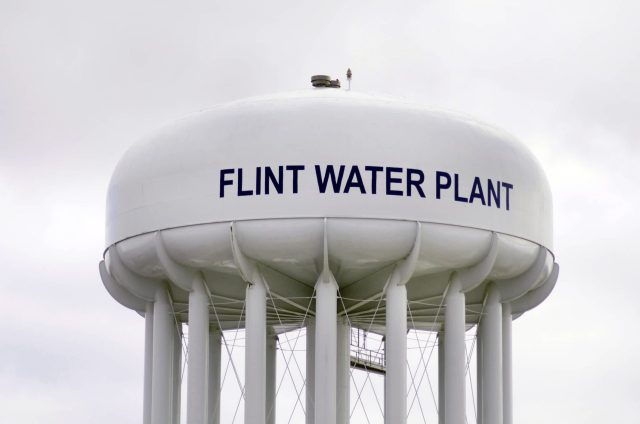 A close-up of a large white water tower labeled "FLINT WATER PLANT" against a cloudy sky.
