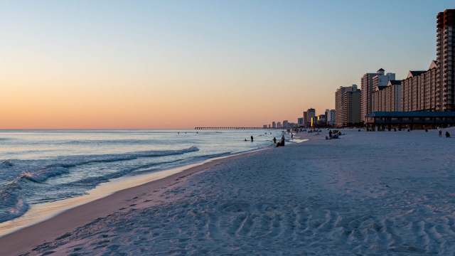 A serene beach scene at sunset with gentle waves, white sand, and tall beachside buildings in the background.