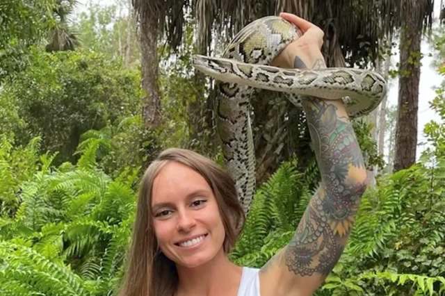 A woman holds a large python while smiling in a lush, green outdoor setting.