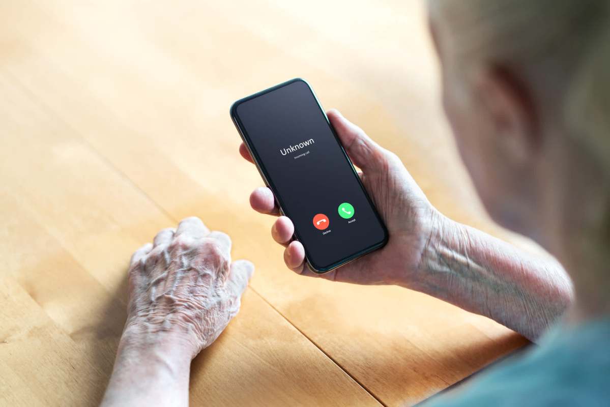 An older adult holds a smartphone displaying an incoming call from "Unknown" on a wooden table.