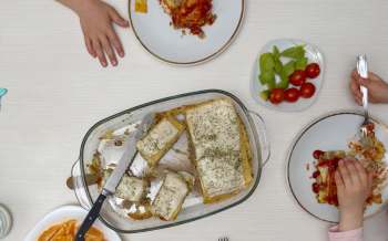 A dinner table scene featuring a dish of baked food, fresh vegetables, and children's hands reaching for the meal.