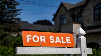 A close-up of an orange "For Sale" sign in front of a large house on a sunny day.