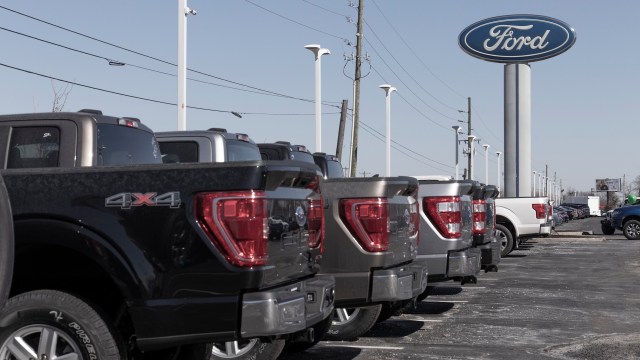 A row of Ford pickup trucks on display in a dealership parking lot with a blue Ford sign in the background.