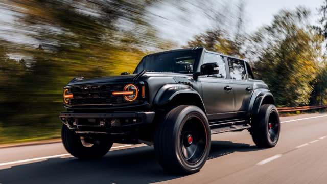 A black Ford Bronco Raptor drives along a highway surrounded by trees.