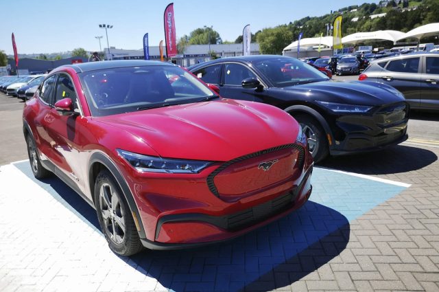Two Ford Mustang electric cars, one red and one black, are parked in a dealership lot under clear blue skies.