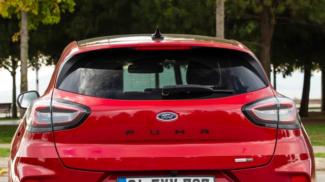 A red Ford Puma parked outdoors, showcasing its rear design and logo among green trees.