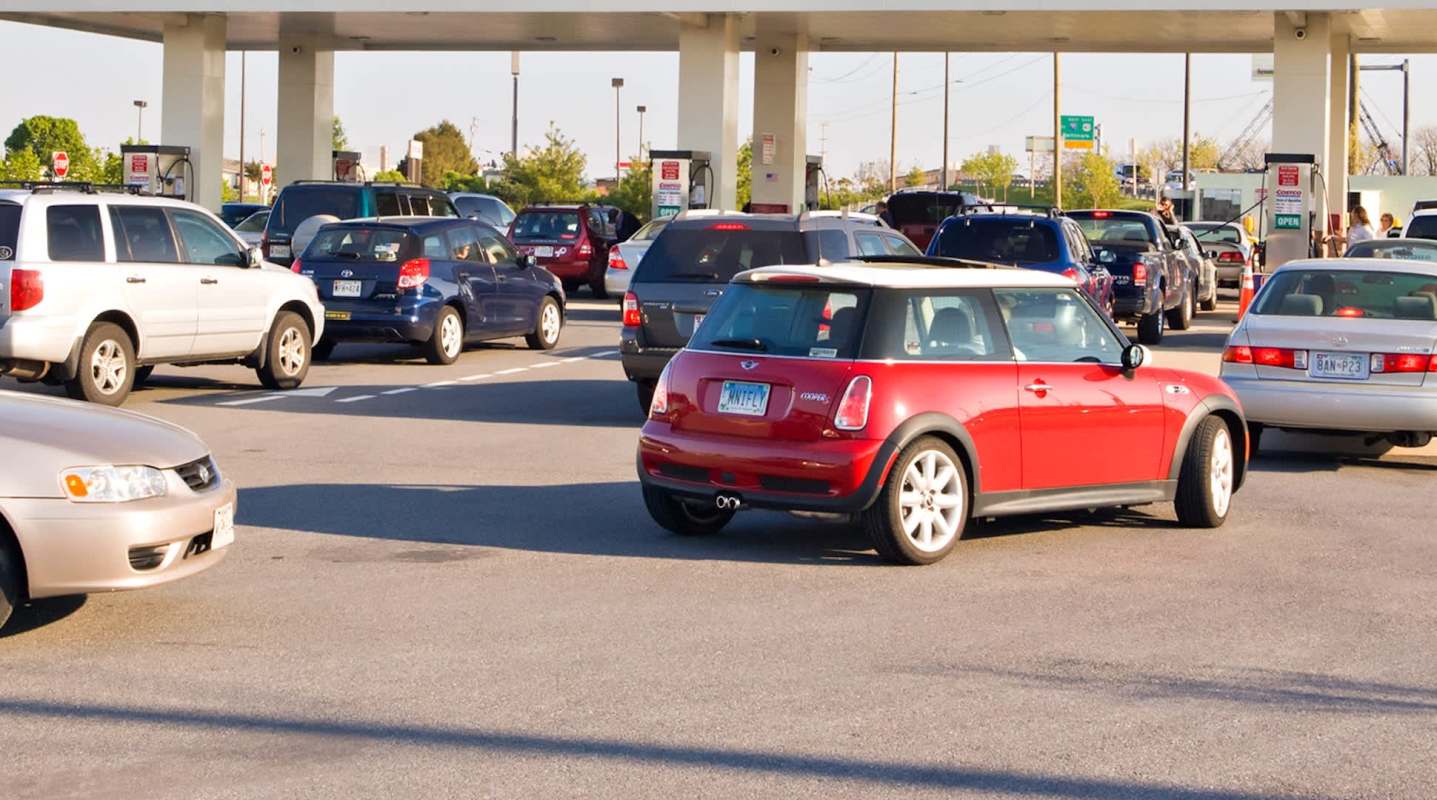 A red Mini Cooper navigates through a busy gas station with numerous vehicles waiting in line.