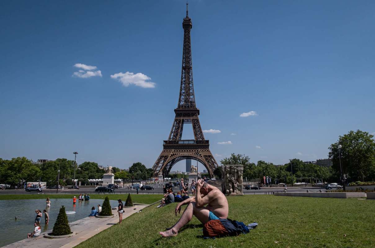 A sunbather sits on grass near the Eiffel Tower, with people and trees visible in the background.