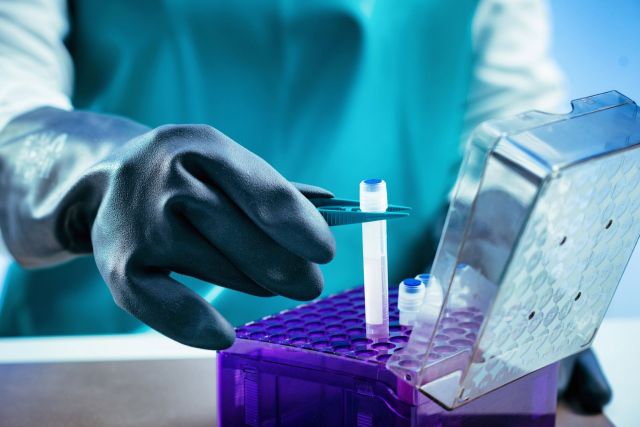 A gloved hand uses tweezers to handle a sample tube in a laboratory setting.