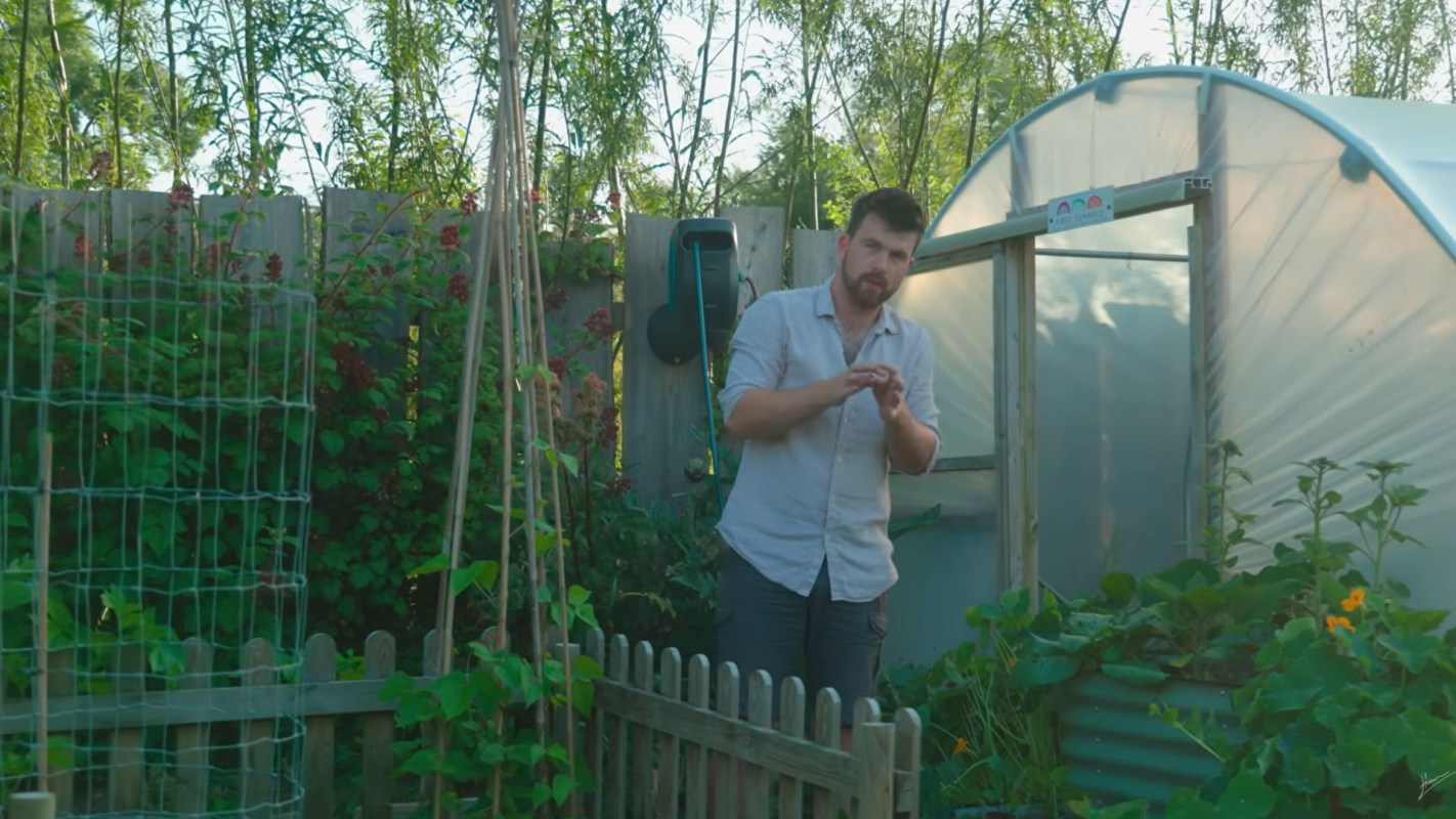 A man standing in a garden outside of a greenhouse.