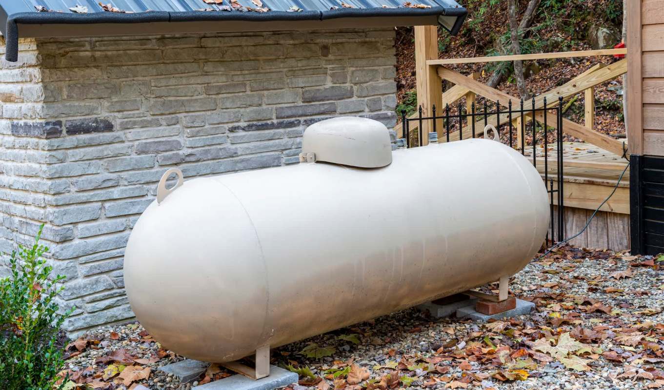 A large beige propane tank sits outside next to a stone wall, surrounded by fallen leaves.