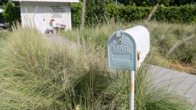 A weathered U.S. mailbox stands amid tall grass near a small white building.
