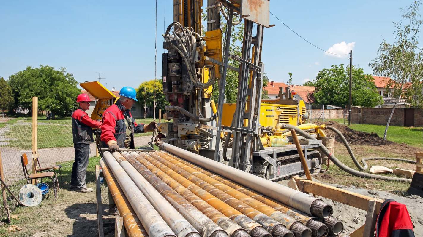 Two workers operate a drilling rig on a construction site, surrounded by equipment and materials under a clear blue sky.