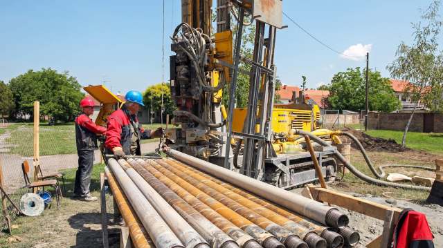 Two workers operate a drilling rig on a construction site, surrounded by equipment and materials under a clear blue sky.