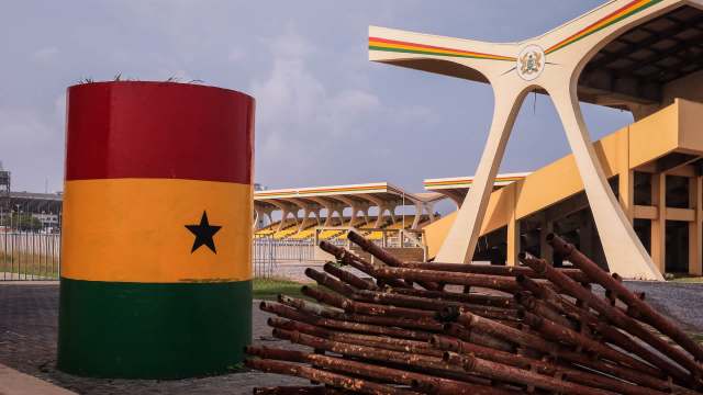 A colorful monument featuring the Ghanaian flag beside stacked rusted pipes at a sports venue.